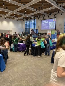 Attendees gather at an indoor fair with various informational tables and a large survey screen.