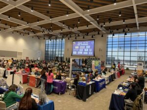 A bustling indoor event features numerous attendees browsing various booths and tables under a sponsor screen. TITLE