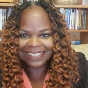 A smiling Black woman with long, curly brown hair and a peach top sits in