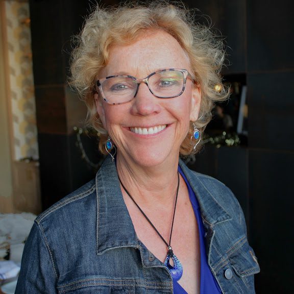 Smiling woman with blonde curly hair, glasses, denim jacket, and blue accessories.