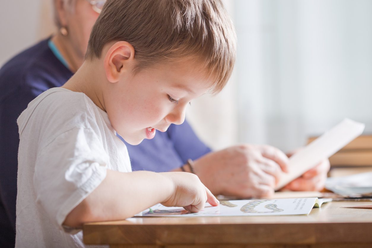 Young boy pointing at a word in his workbook while an adult sits beside him at the table