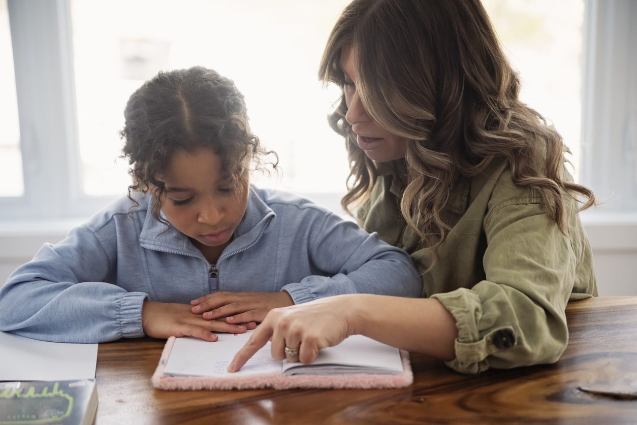 Mother sitting beside her young daughter at a dining table, pointing to a page in her notebook