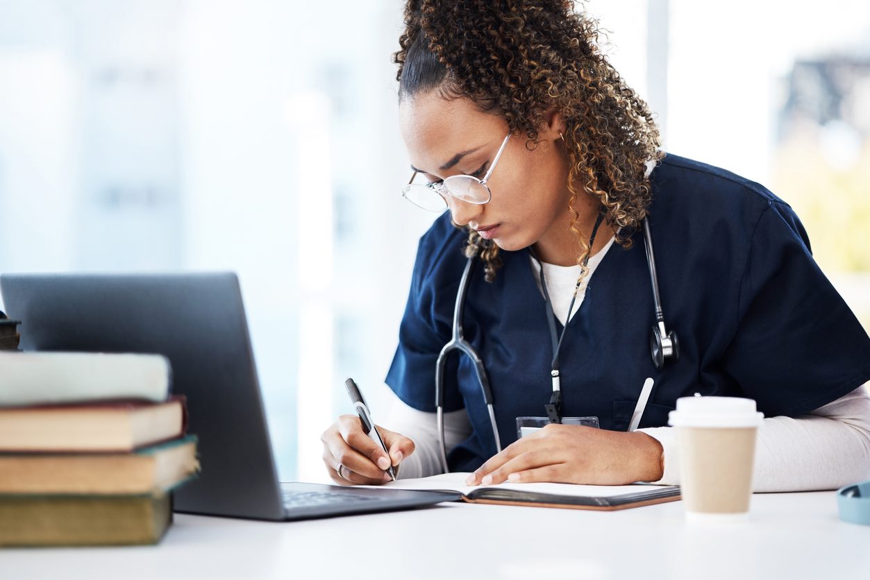 Nursing student writing notes at a desk with a laptop and stacked textbooks