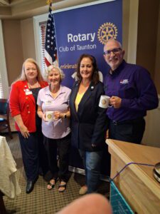 Four smiling people, two holding Rotary mugs, pose before a Rotary Club of Taunton banner