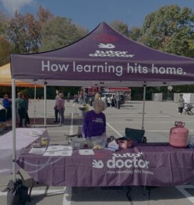 A smiling woman in a purple Tutor Doctor shirt stands at a branded outdoor event booth.