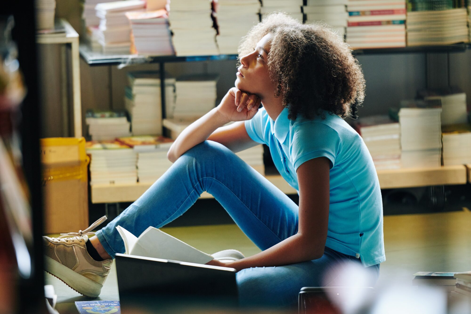 A student sitting on the floor of a library, looking up in thought with a book open in her lap