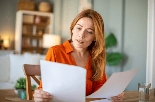 A focused woman with red hair reviews documents at a table in a home setting. TITLE: