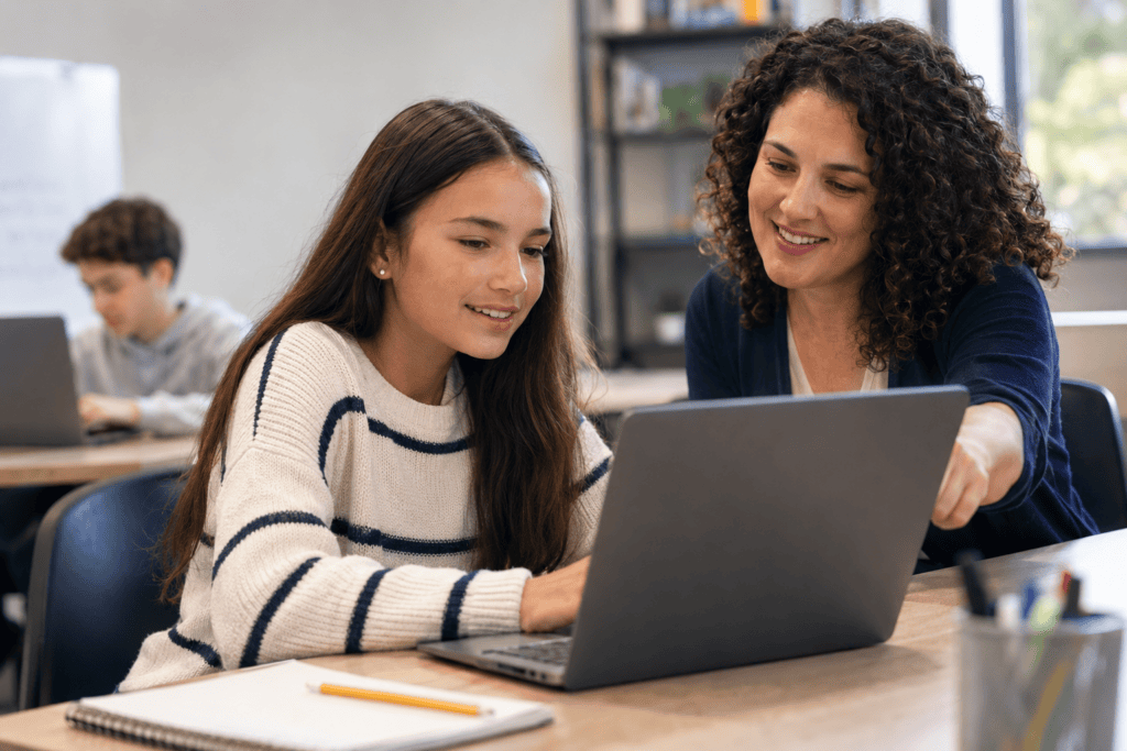 A smiling teacher guides a student working on a laptop in a bright classroom.